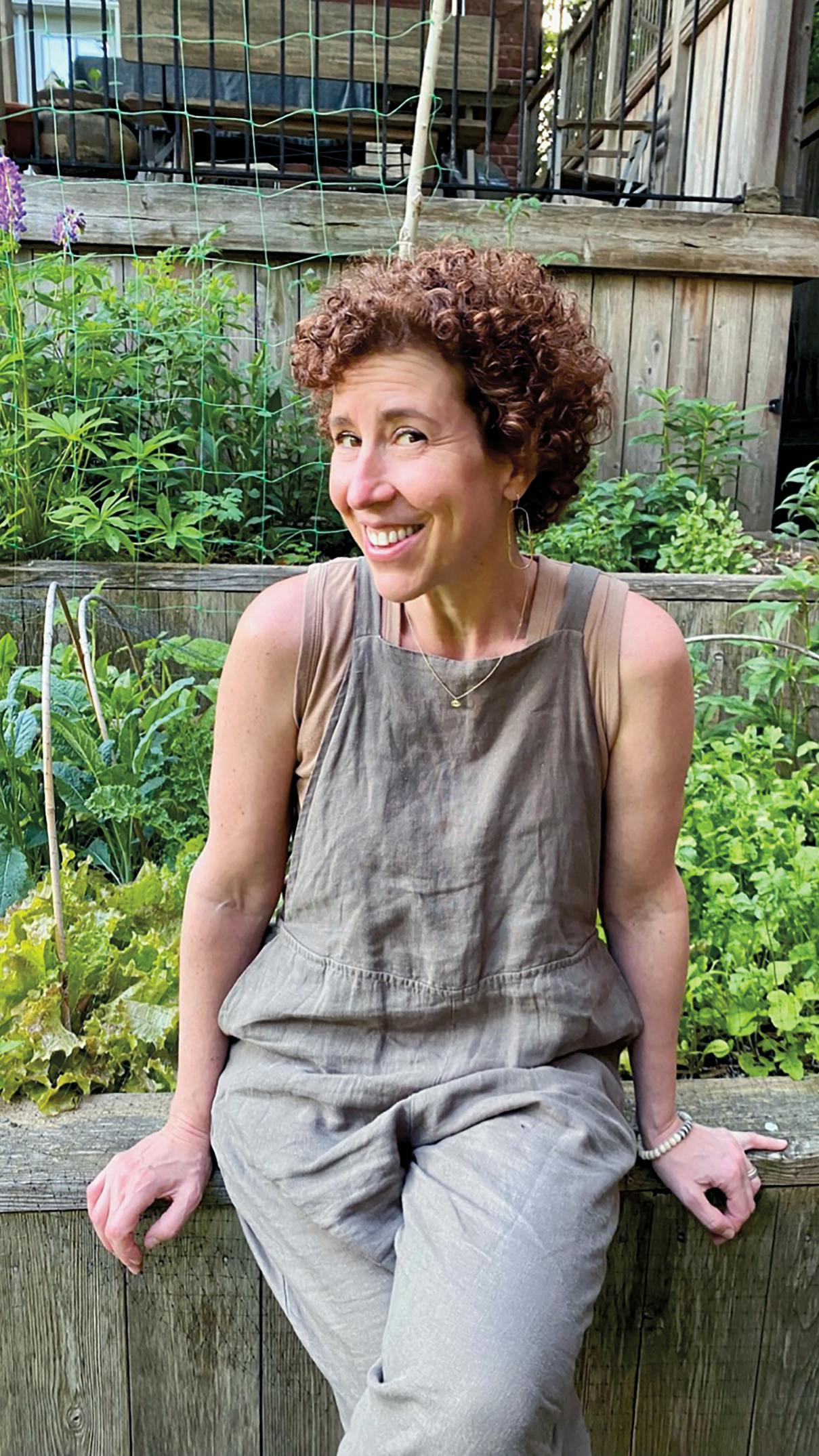 Person with curly brown hair in gray overalls sitting on wooden ledge in vegetable garden
