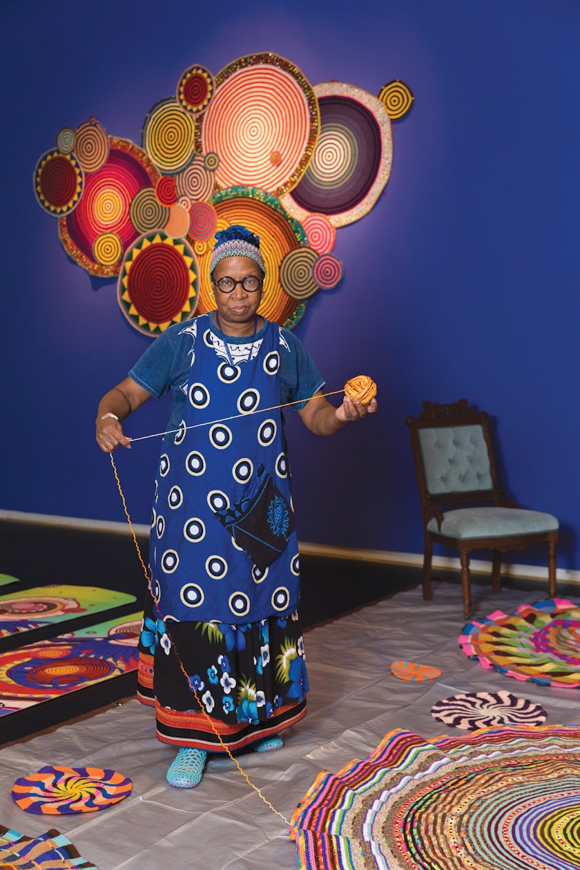 Artist creating large colorful crochet artwork with finished work on blue wall in background