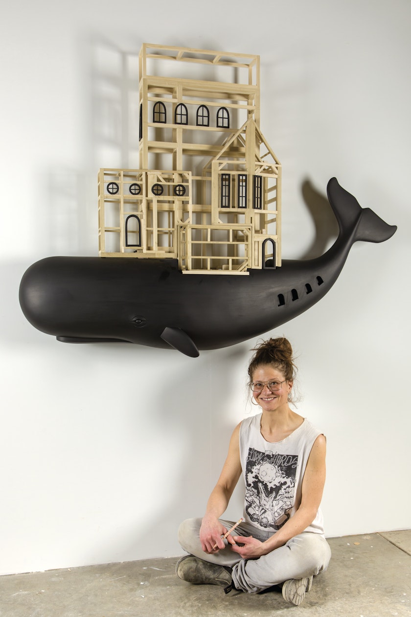 Artist sitting beneath a wood sculpture of a black whale with a building on its back