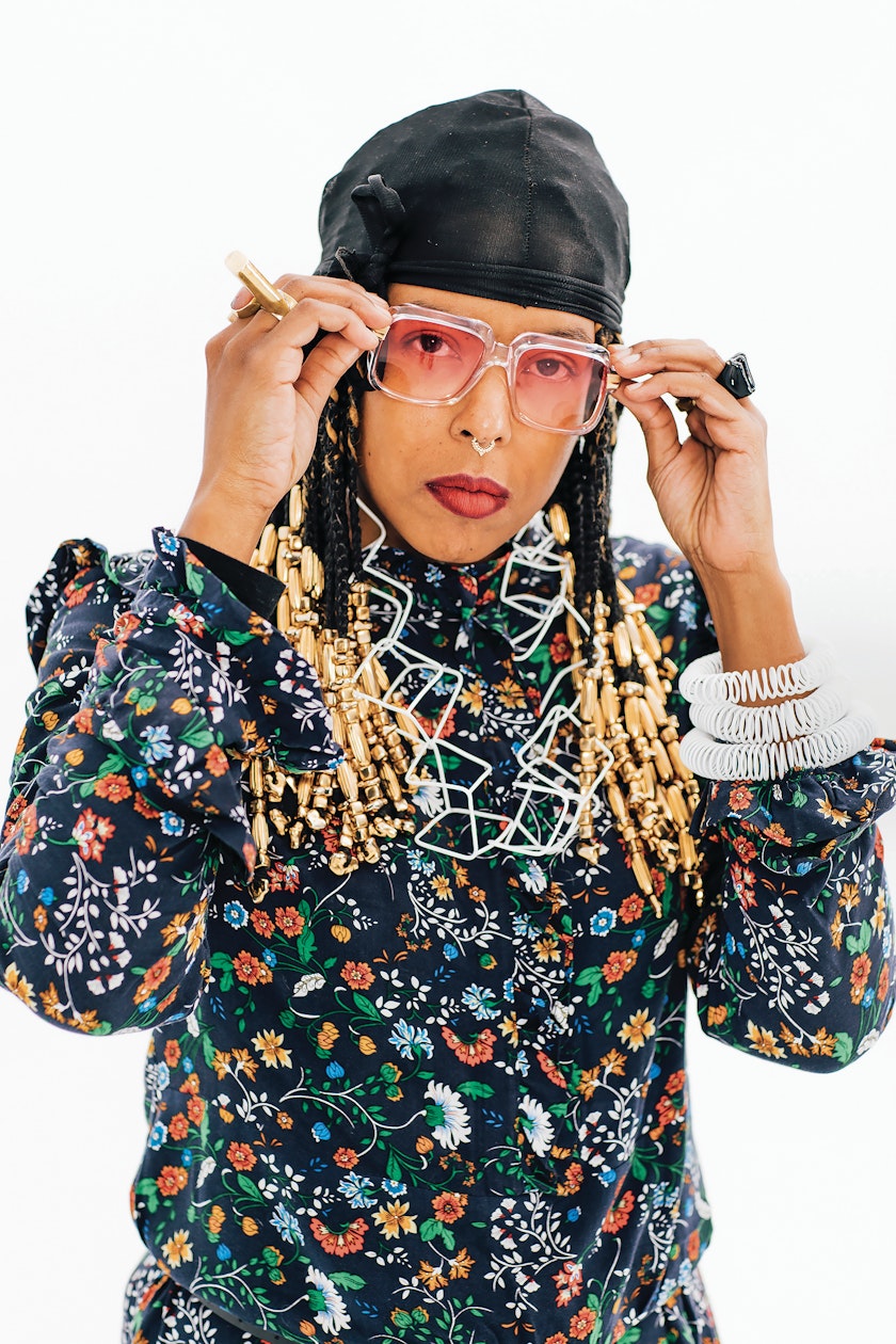 Portrait of Tiff Massey in floral patterned shirt and black cap white necklace and bracelet adjusting pink shaded sunglasses