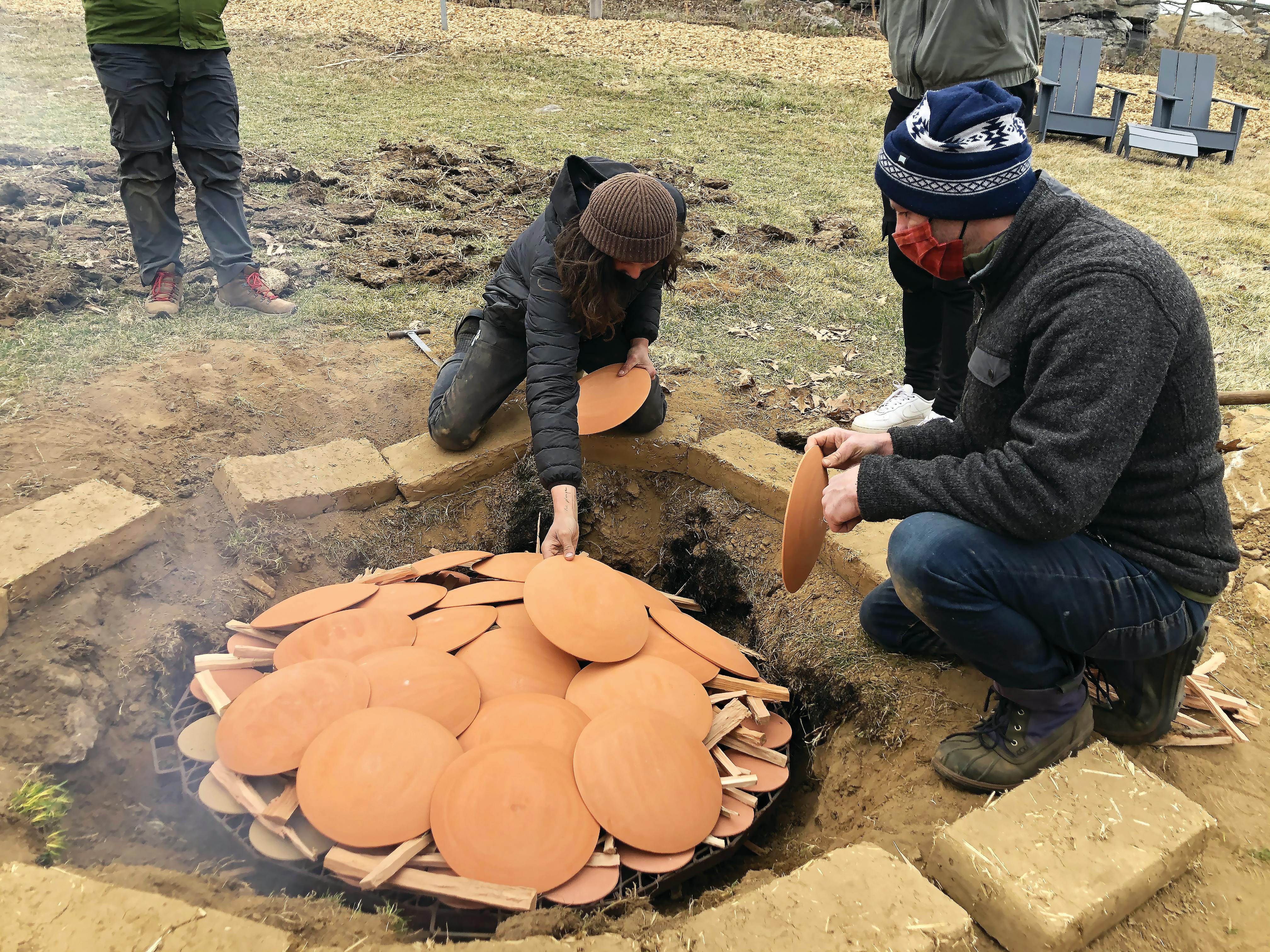 two crouched people in jackets hats and masks stacking clay plates over wood to prepare a buried pit fire