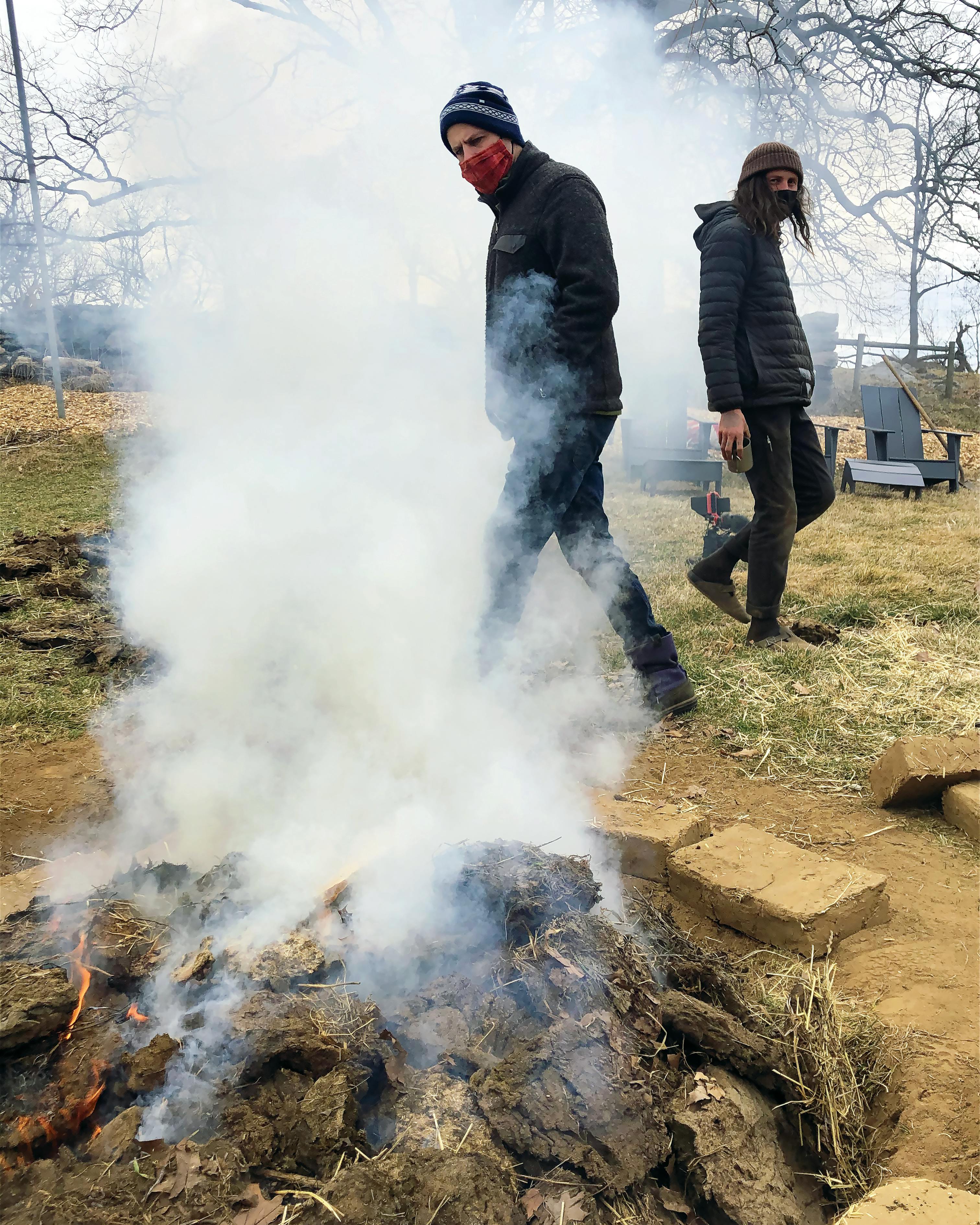 two masked people walking around a smoking pit fire