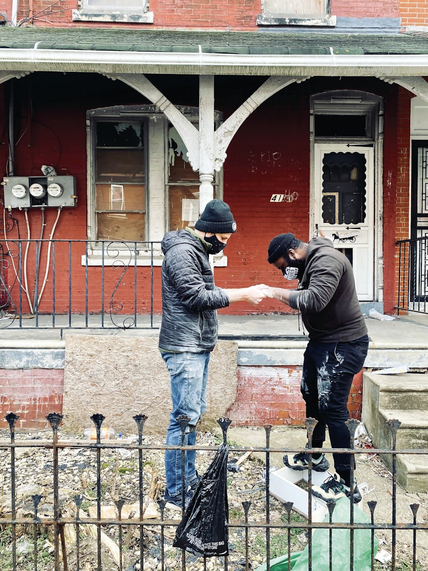 Two people making a plaster cast outside in a yard in front of a red brick house with boarded windows on what appears to be a chilly day
