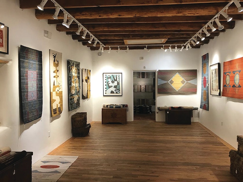 view of the inside of a small art gallery with wooden floor and cream walls and weavings and paintings on display