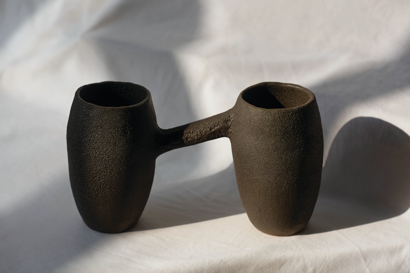 brown ceramic piece with two vessels connected by a thin slanted bar arranged against a gray background