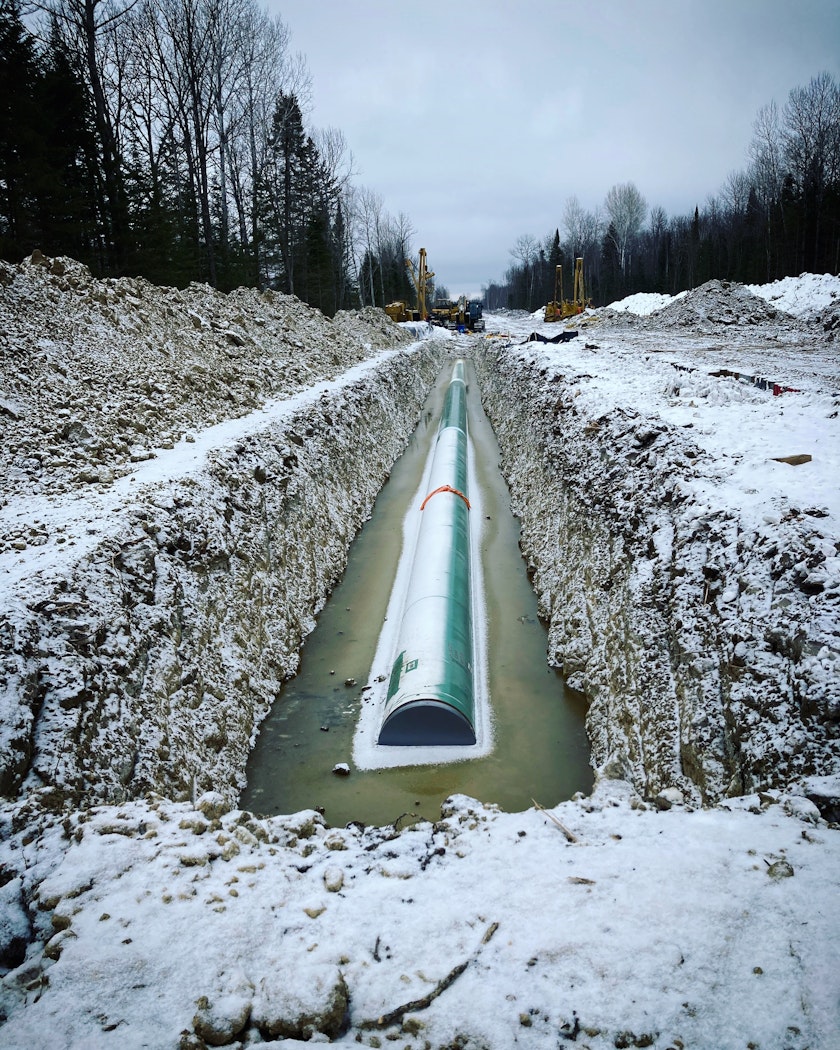 A pipeline construction site in winter flanked by a forest. In a deep ditch in the snowy earth lies a large frosty pipe half submerged in murky water.