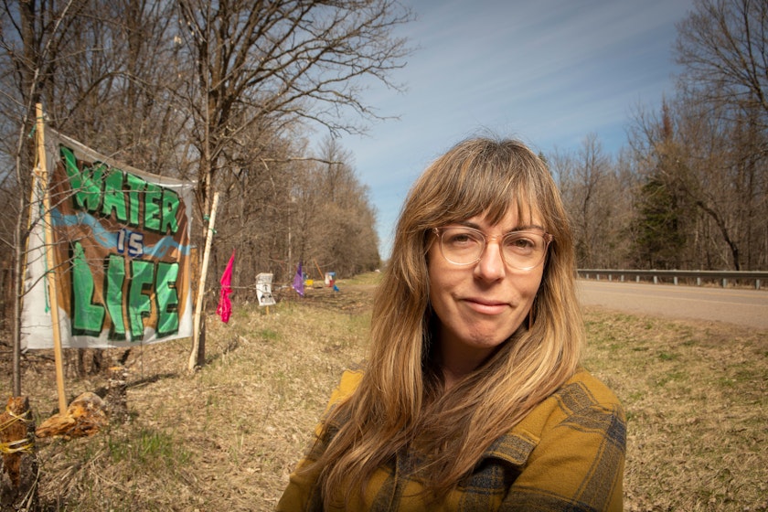 Portrait of a woman in a plaid shirt standing in the ditch beside a rural road with a handmade fabric sign hanging against the treeline that says water is life