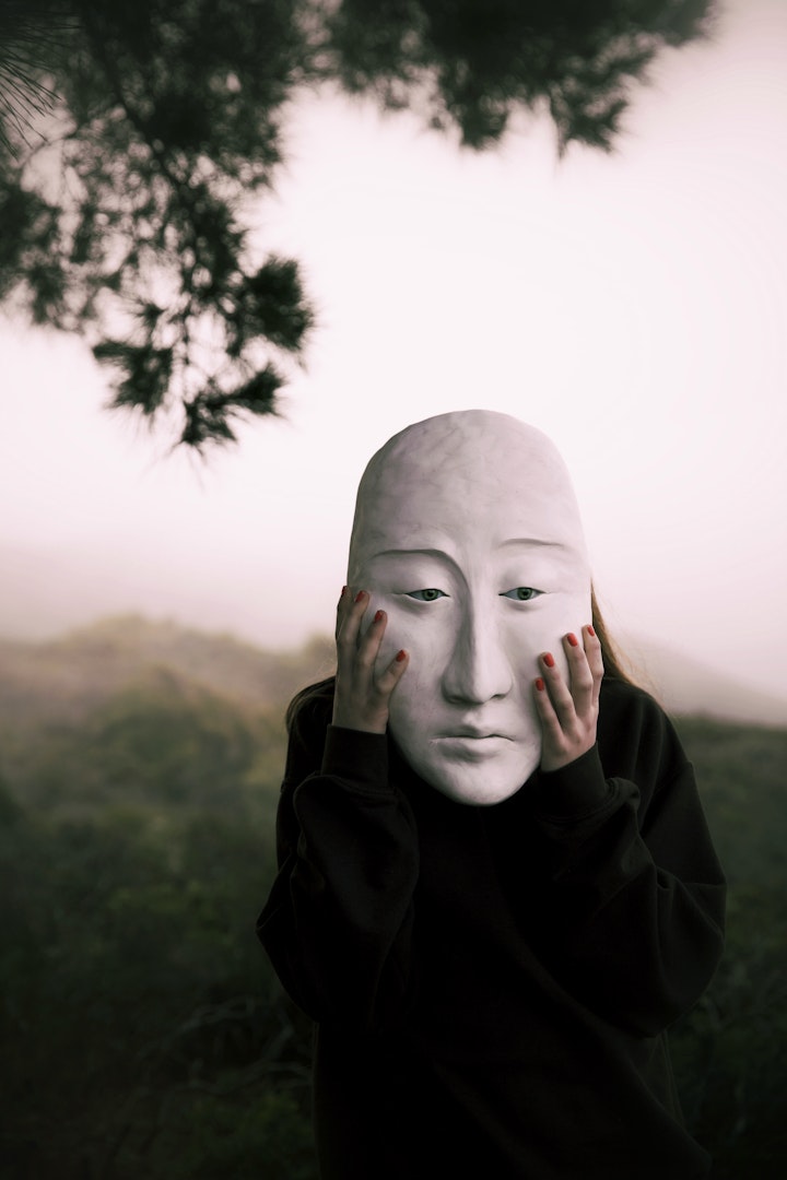 photo of a model holding up a large white contemplative paper mache mask with blue glass eyes
