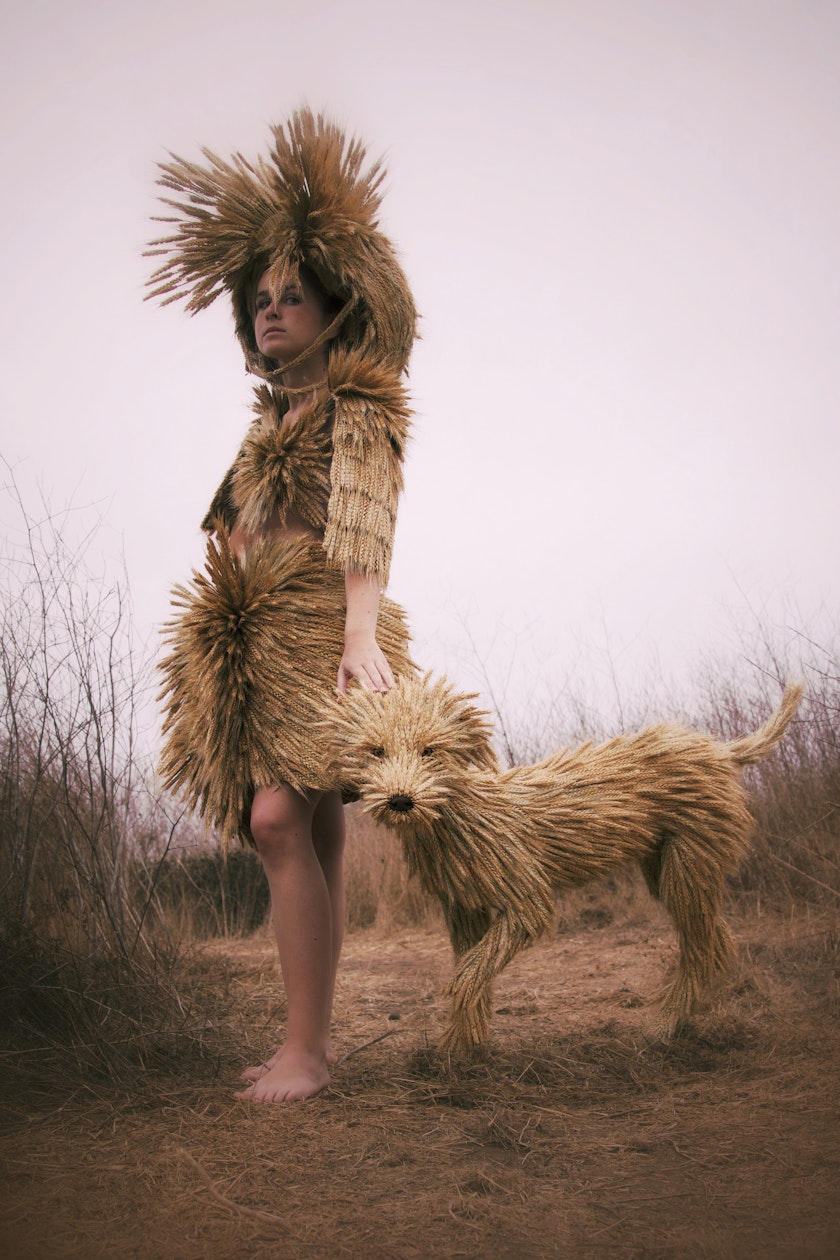 model wearing sculptural headpiece top and skirt made from heads of wheat posing beside a sculpture of a dog also made from wheat