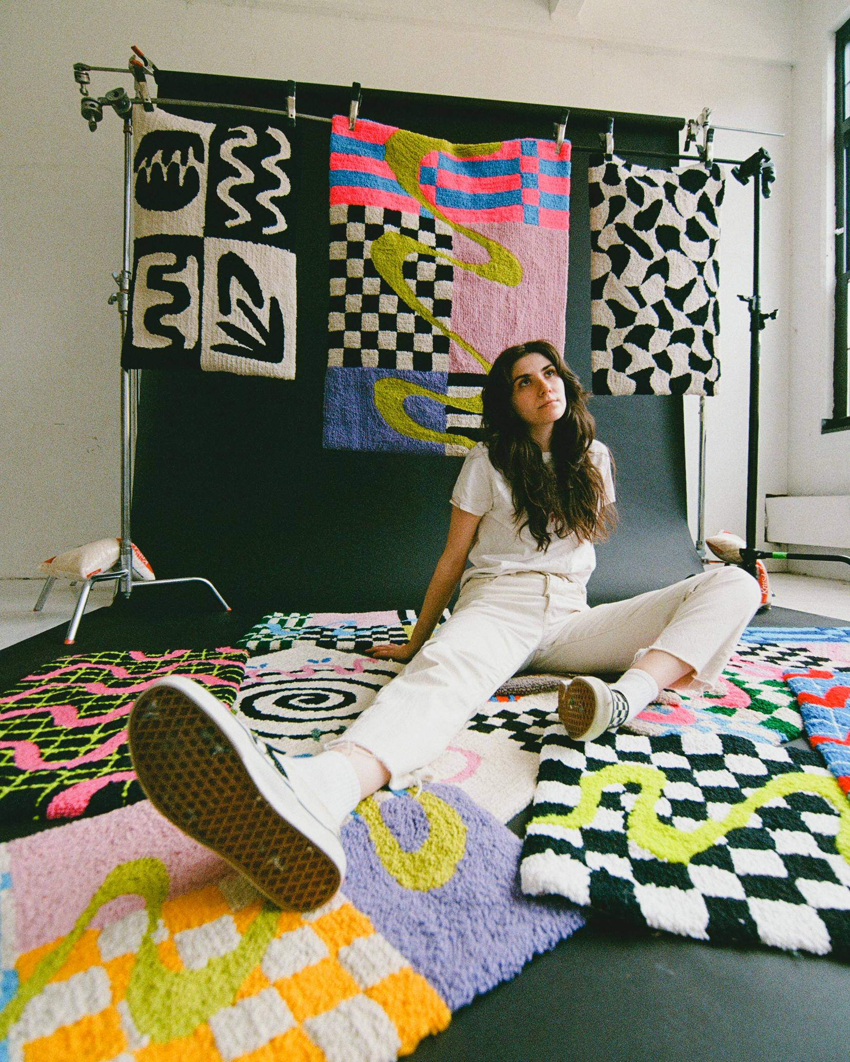 artist posing on floor with arrangement of colorful hand tufted rugs in displayed in studio against a black backdrop