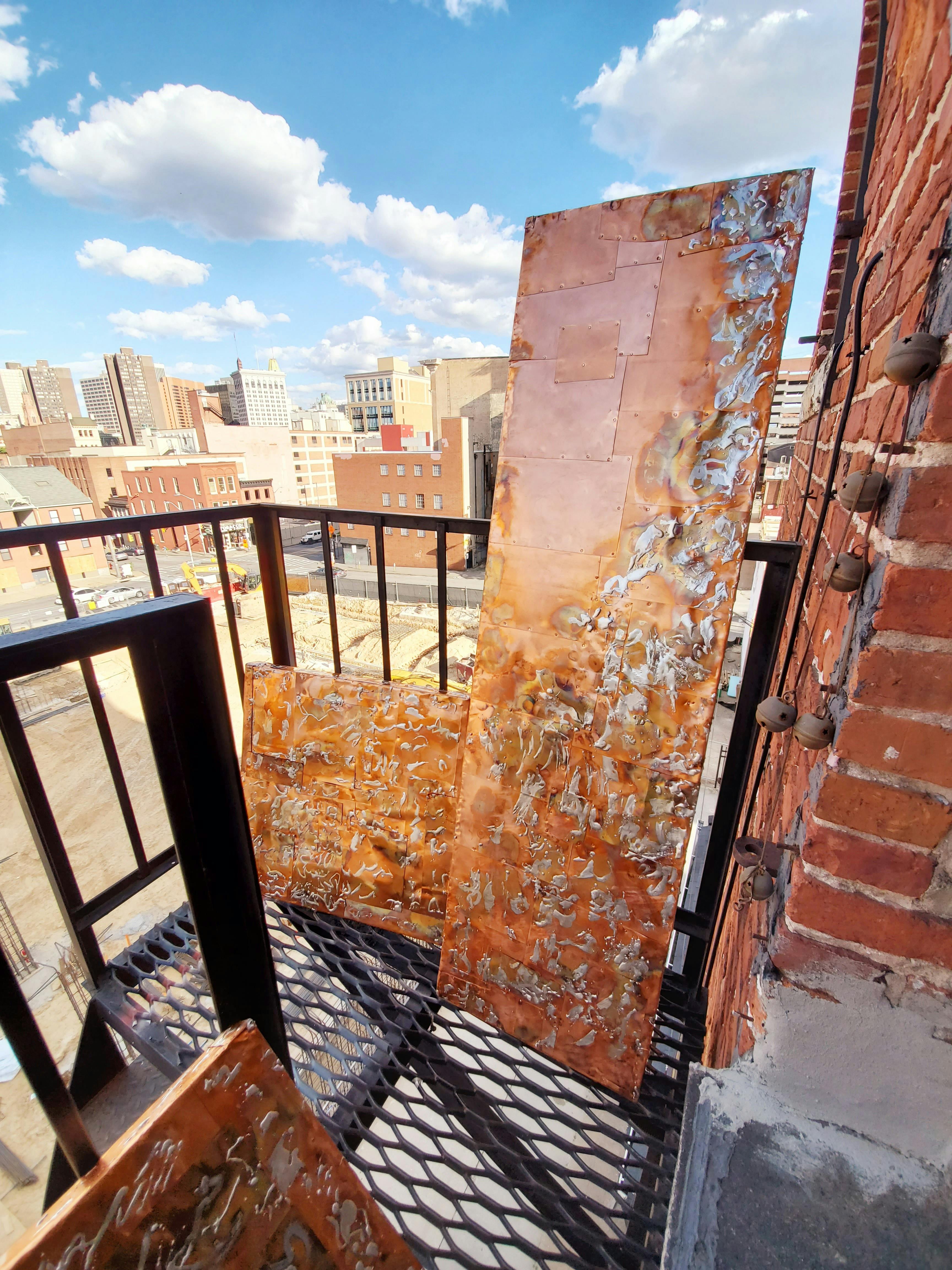 series of rectangule wallhangings featuring textured copper arranged on fire escape overlooking a city on a sunny day