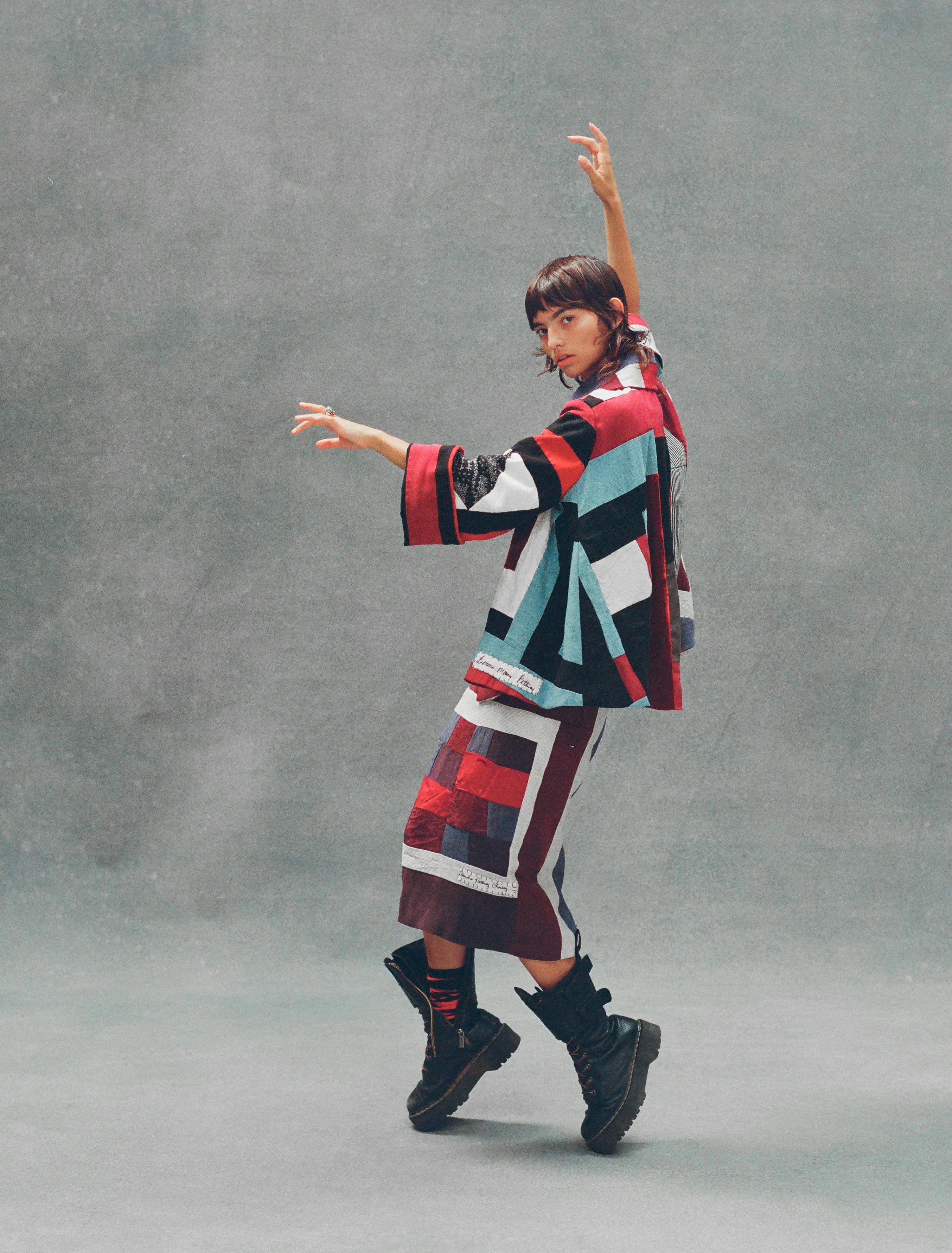 model in dancing pose wearing large black lugsole boots and pathwork quilt top and skirt with red black and blue 