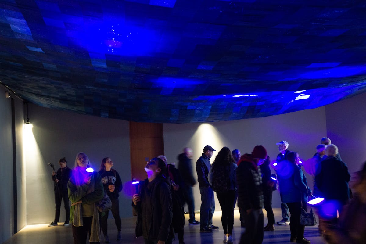 gallery space filled with people shining flashlights up onto a dark blue pathwork fabric draped across the ceiling