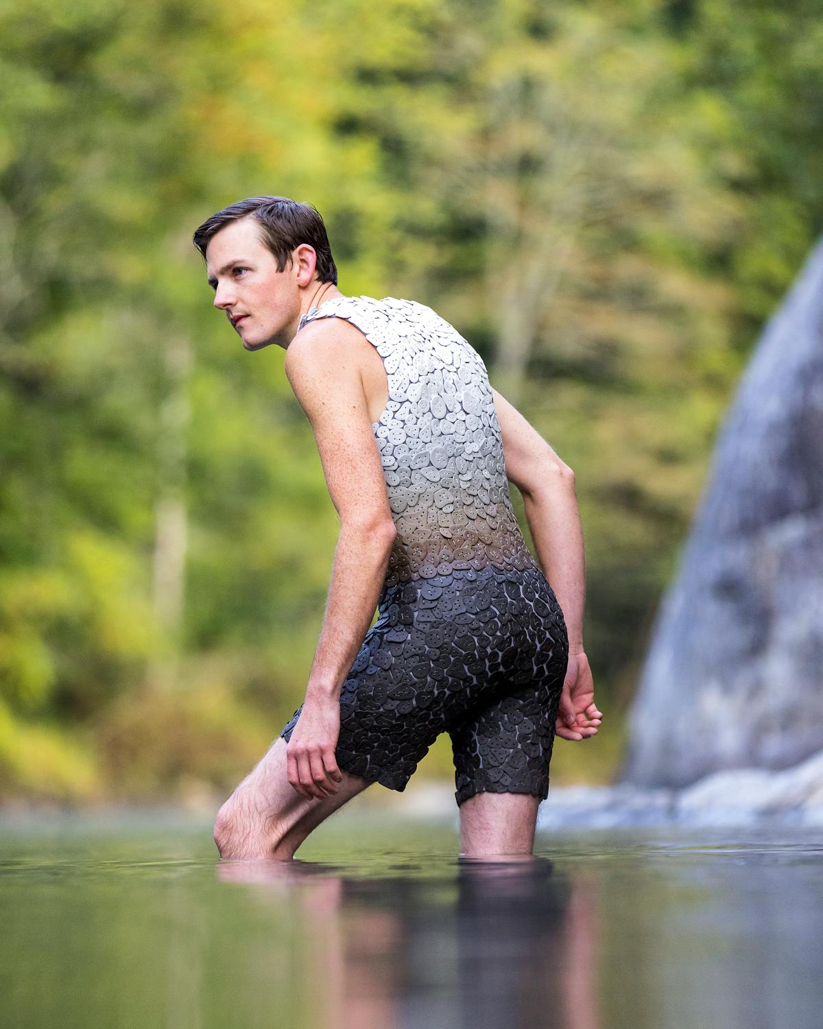 man wearing a one-peice swimsuit made from ceramic tiles in a gradient of brown up to cream while wading into a river