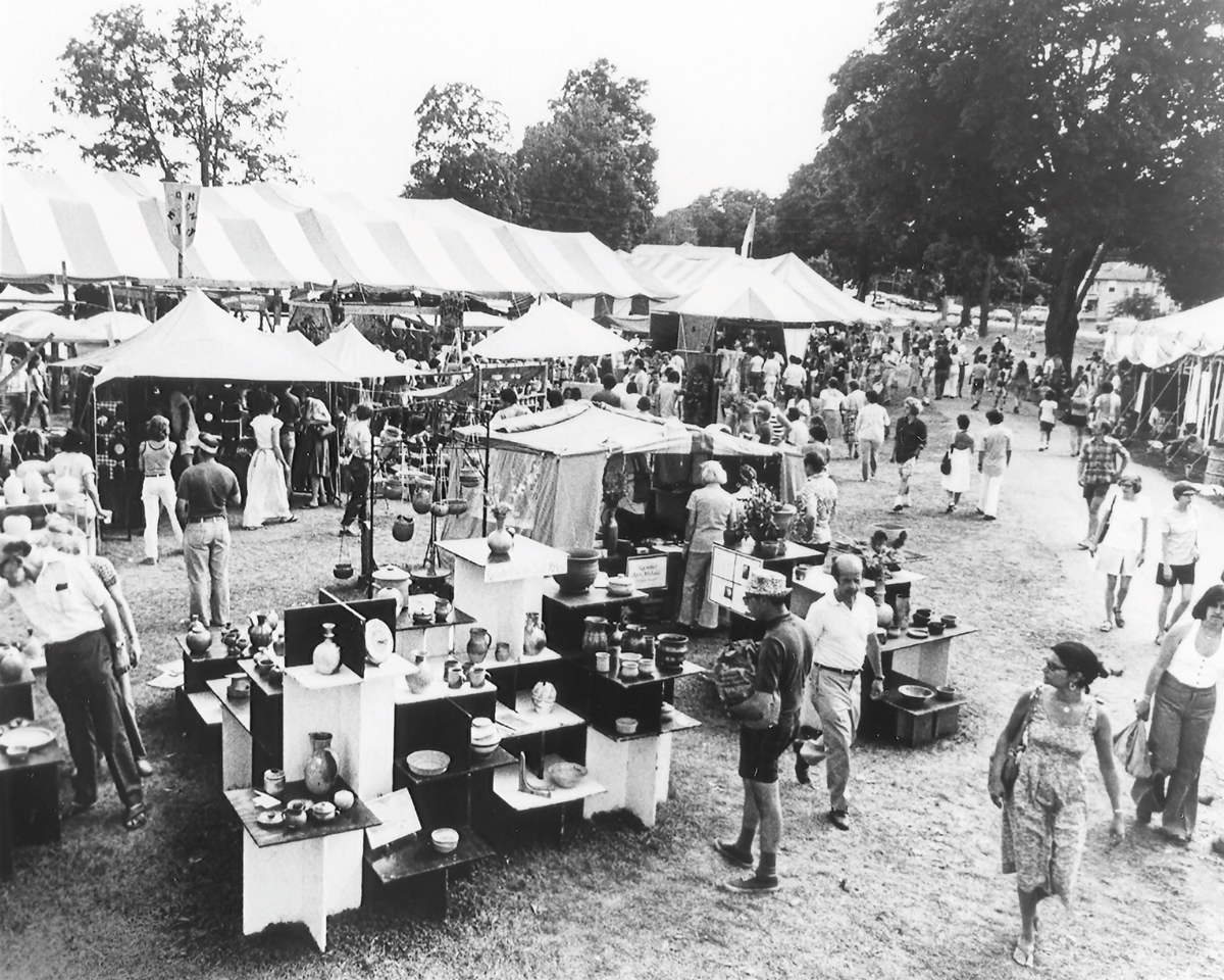 black and white photo of and outdoor craft fair with visitors walking amid displays of work