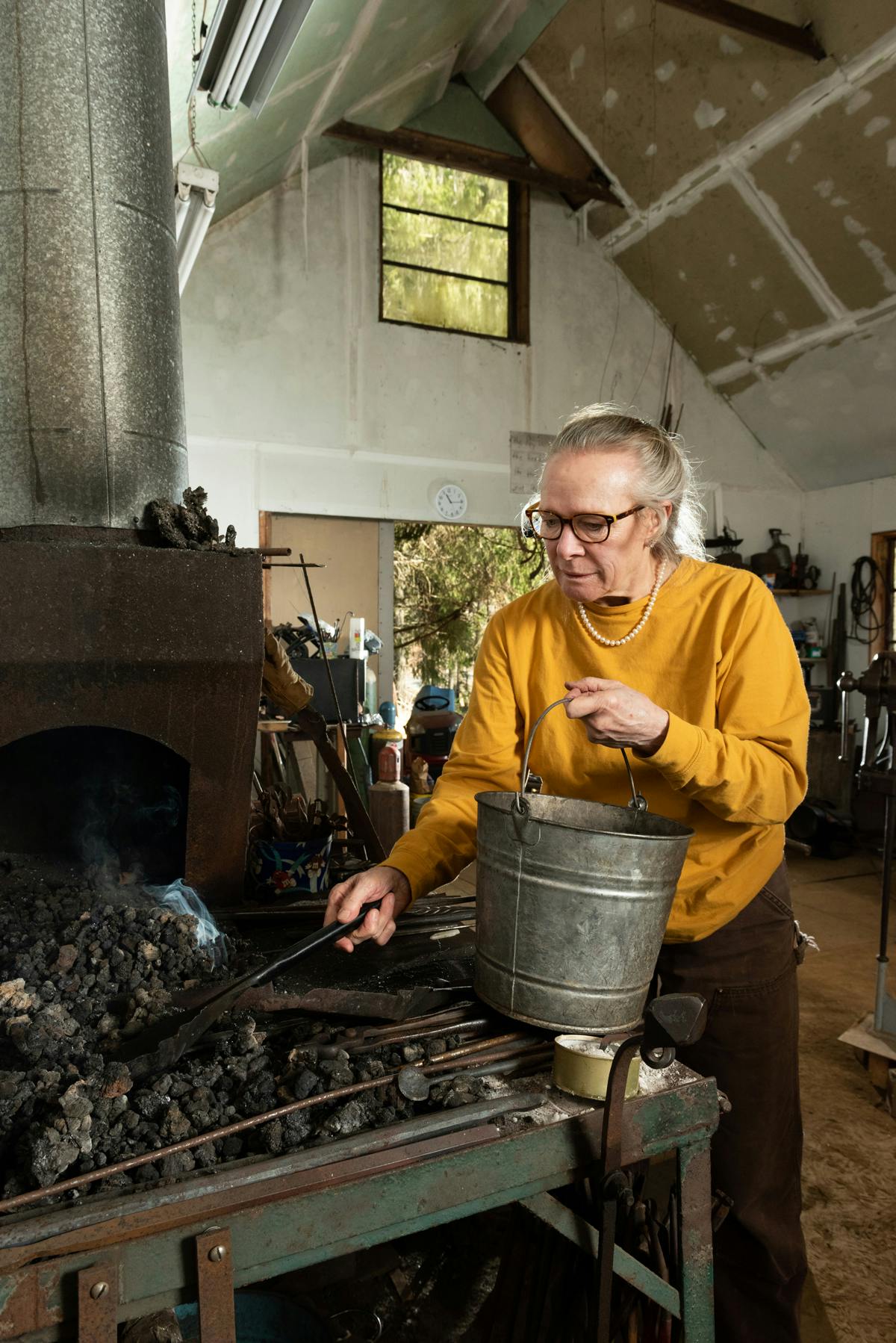 photo of blacksmith wearing yellow sweater and pearls shoveling coal into a bucket