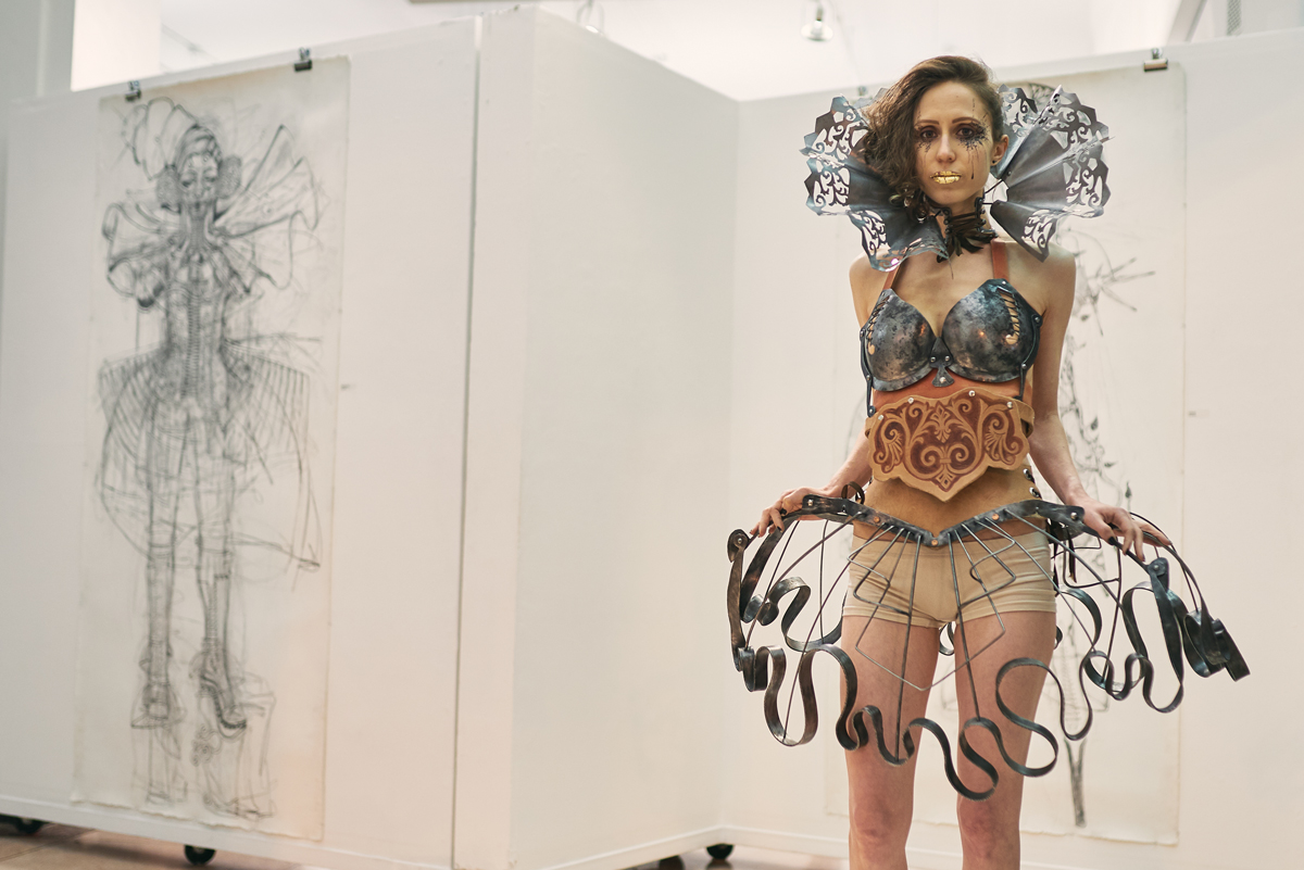 model in exhibition space wearing fashion attire that's part burlesque part leather armor and part wrought iron ornamentation