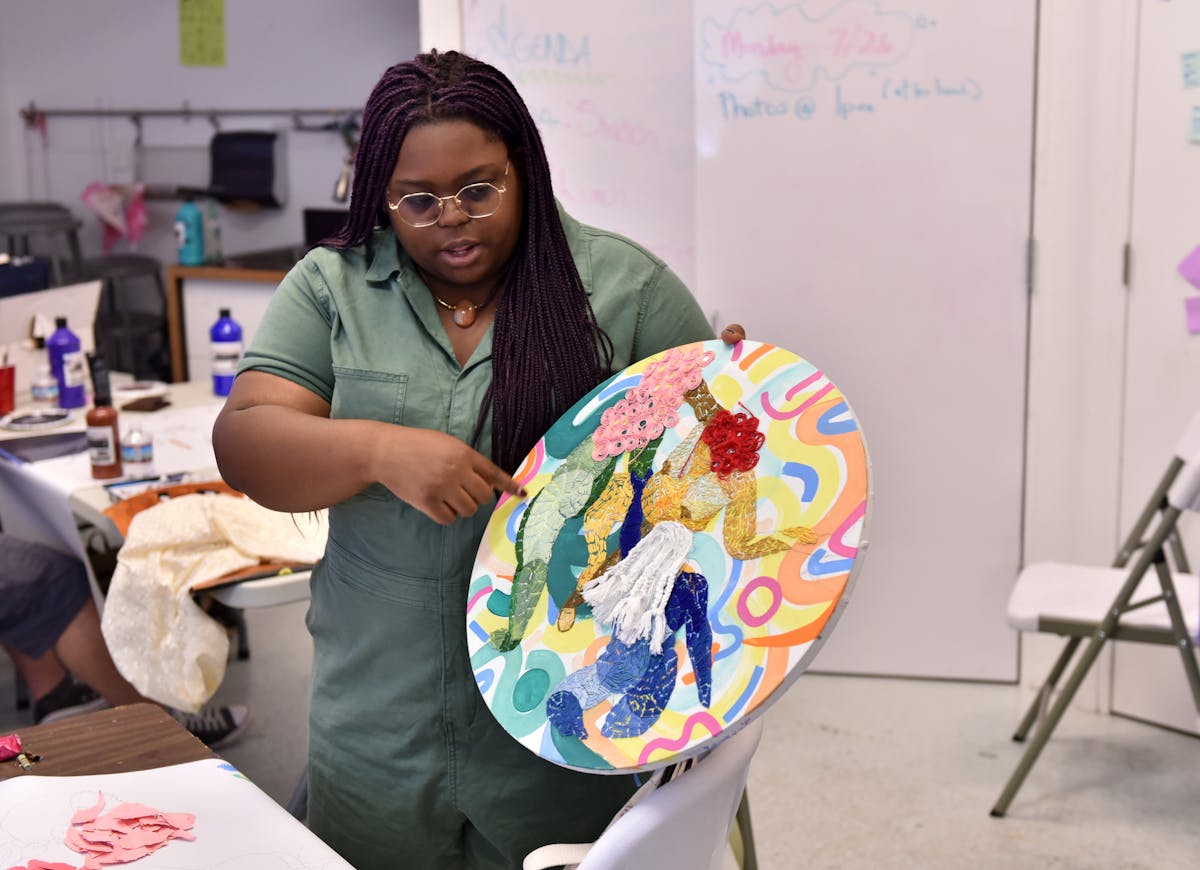artist holding up and pointing at a round painting with embroidered design in classroom setting