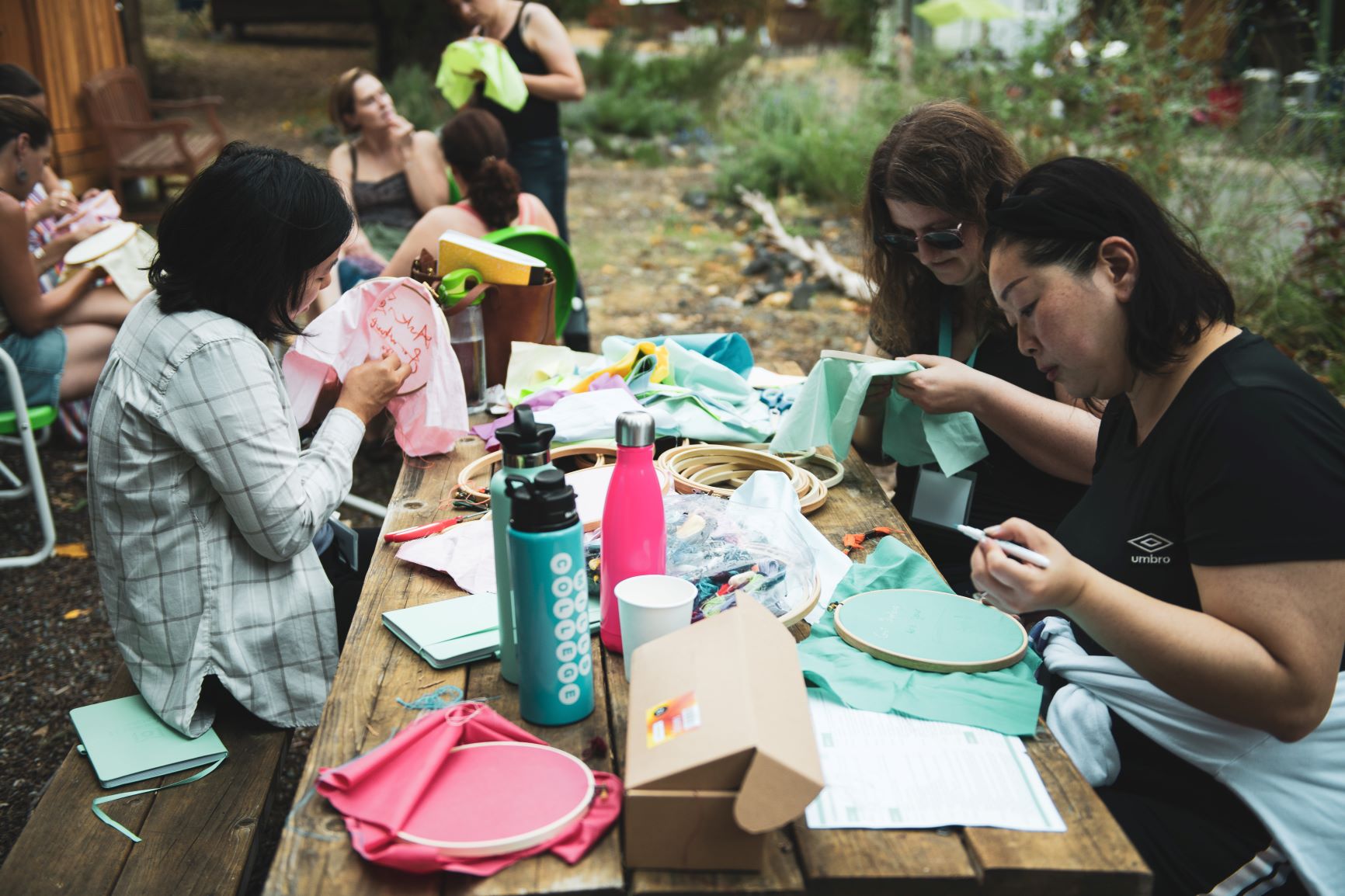 group of people sitting around a table outdoors working on embroidery projects