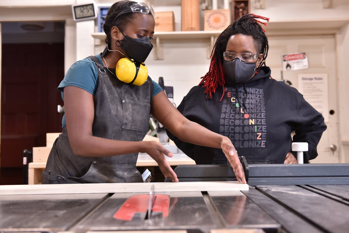two women wearing face masks and protective glasses working in a shop at a table saw