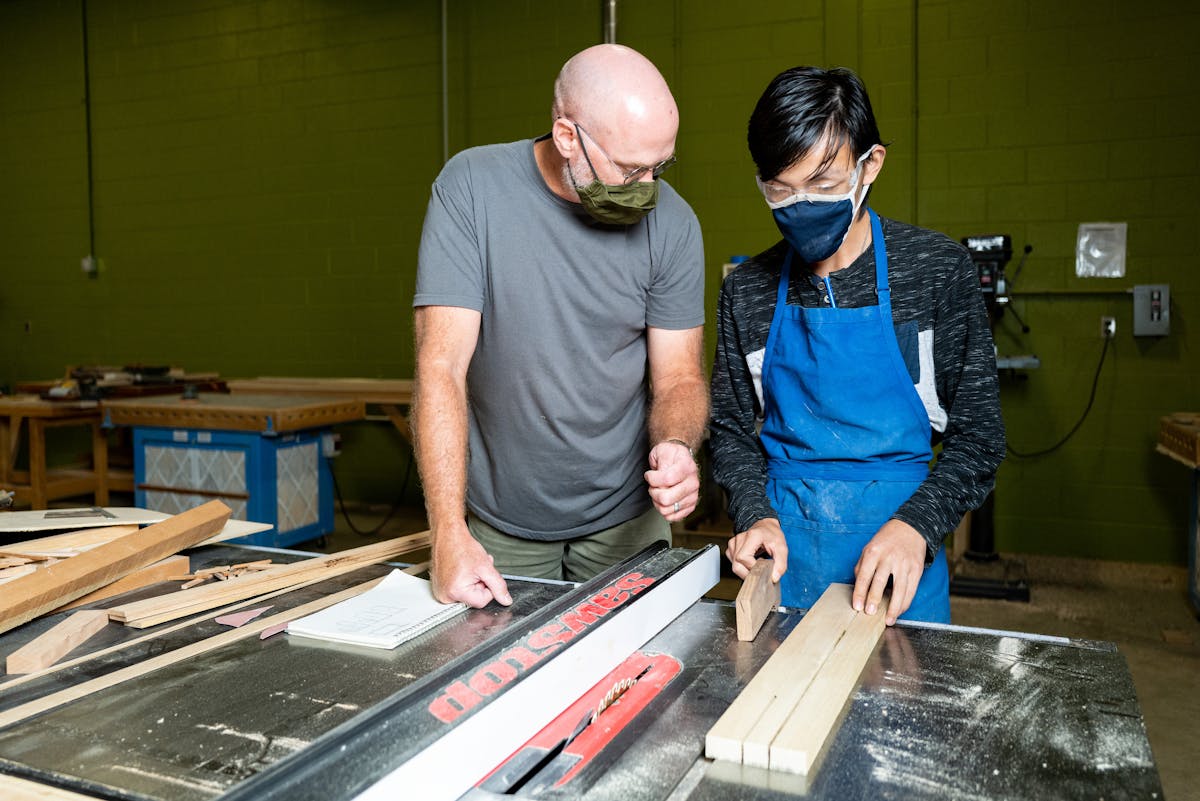 teacher and student wearing masks and working at table saw in a workshop