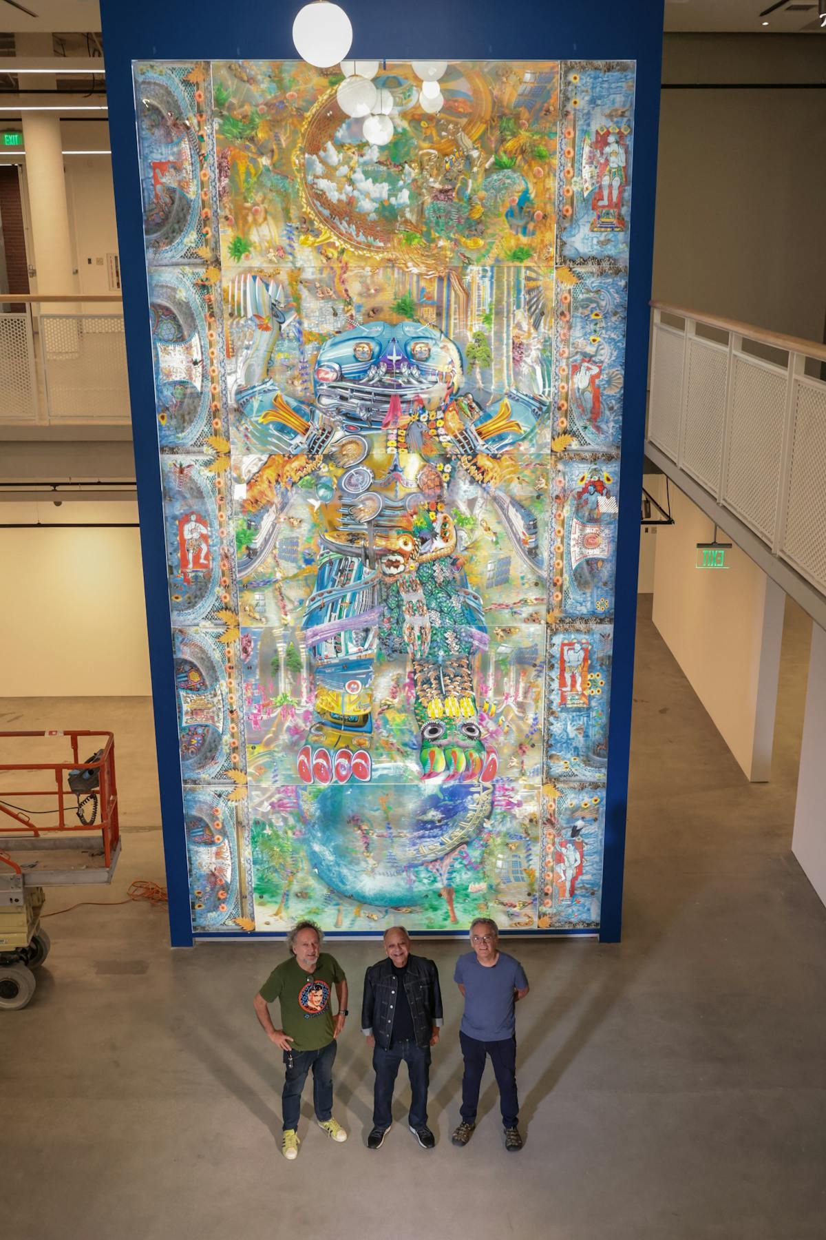 three people standing in front of a large colorful floor to ceiling artwork depicting a figure at once psychedelic and robotlike