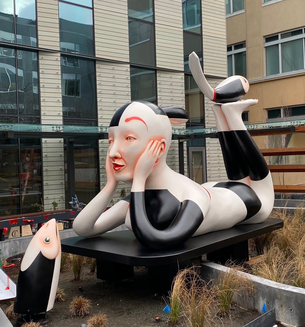 large sculptural installation in a courtyard showing a black and white patterned human figure on their belly with feet up and a fish head coming out of the ground