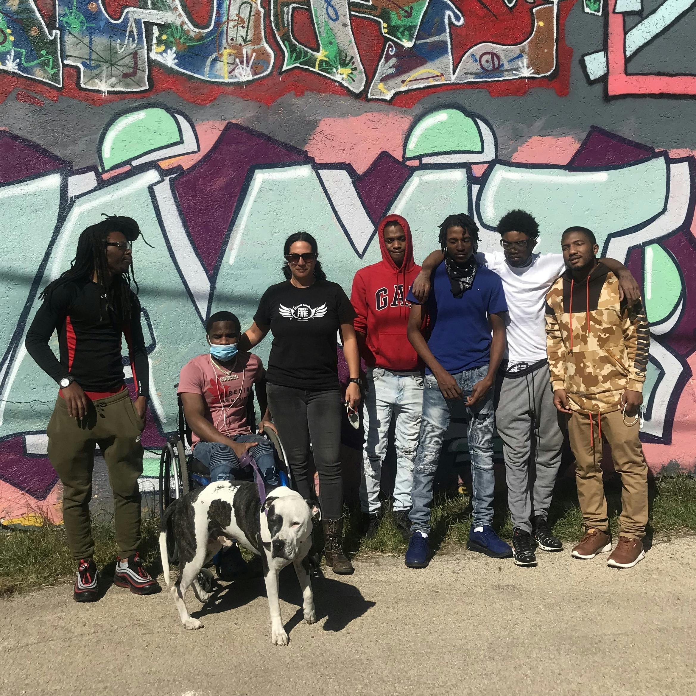 Row of people posed in front of a graffitied wall with a pitbull