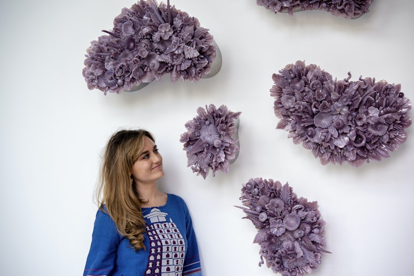 portrait of artist looking up at a series o wall sculptures that look like bunches of flower pedals made from purple glass all hanging on a white wall