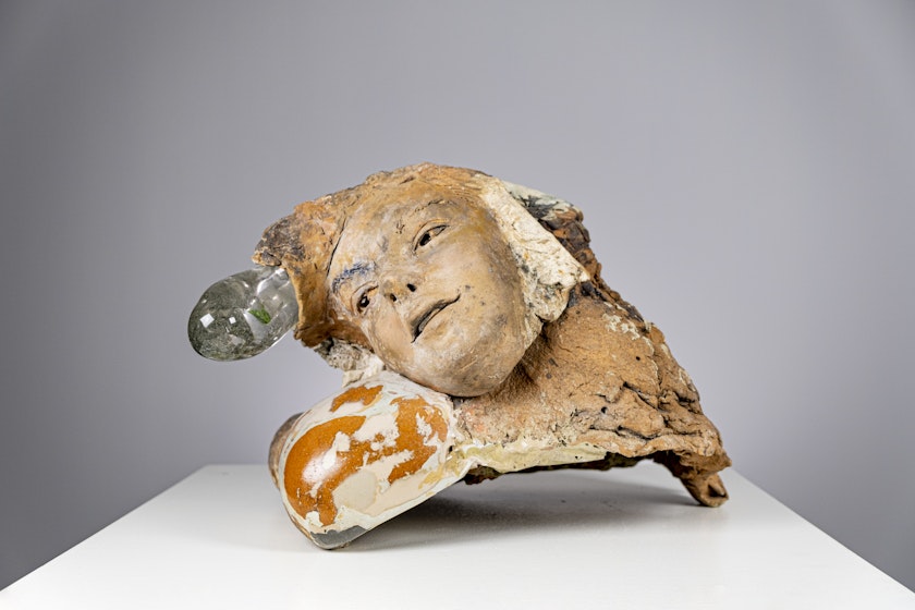 ceramic sculpture of weathered tan face looking askew and melded with amorphous clay and glass shapes on a white table gray background