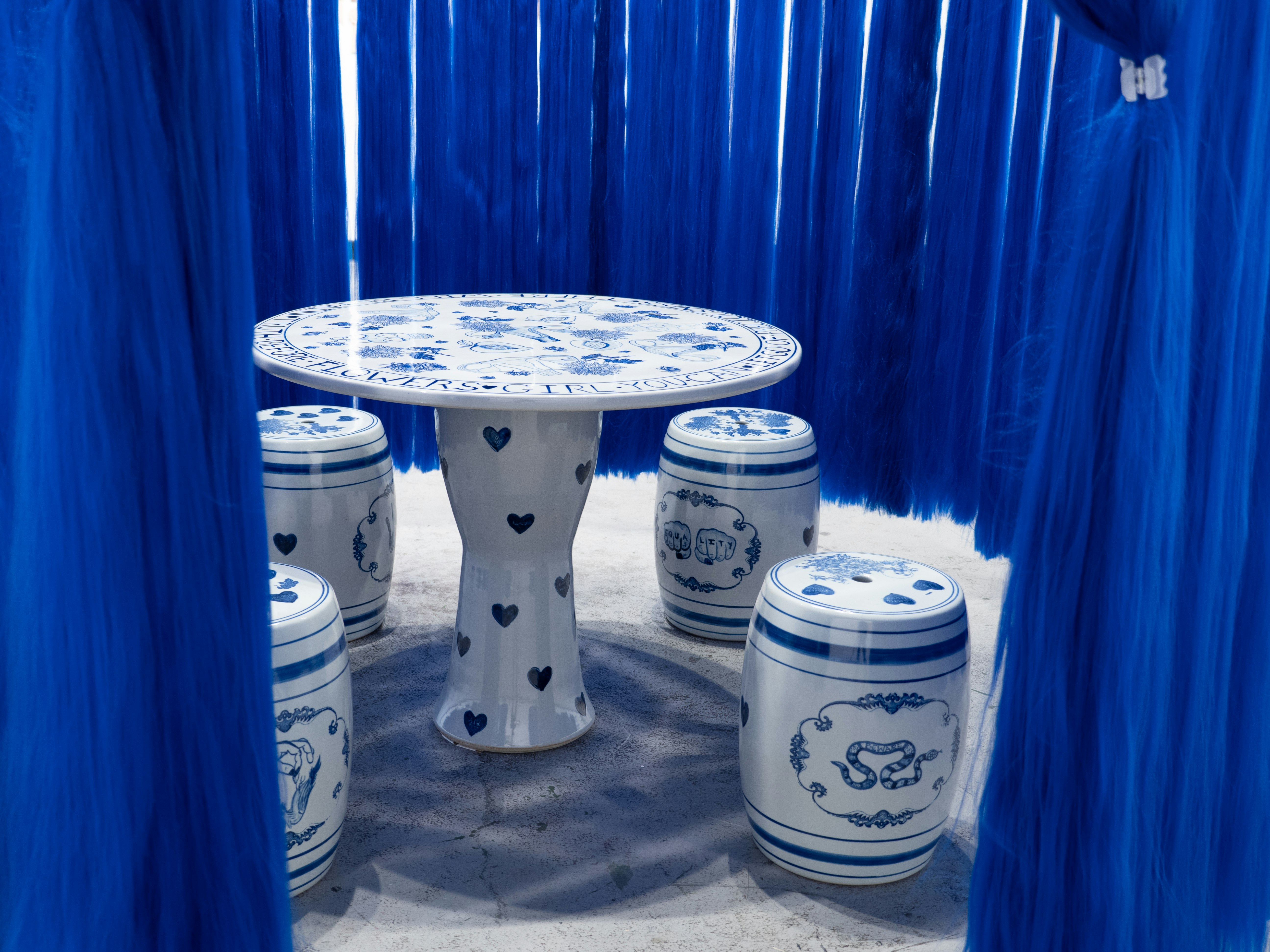 white porcelain table and with four barrel shaped stools with blue designs surrounded by a curtain of blue hair