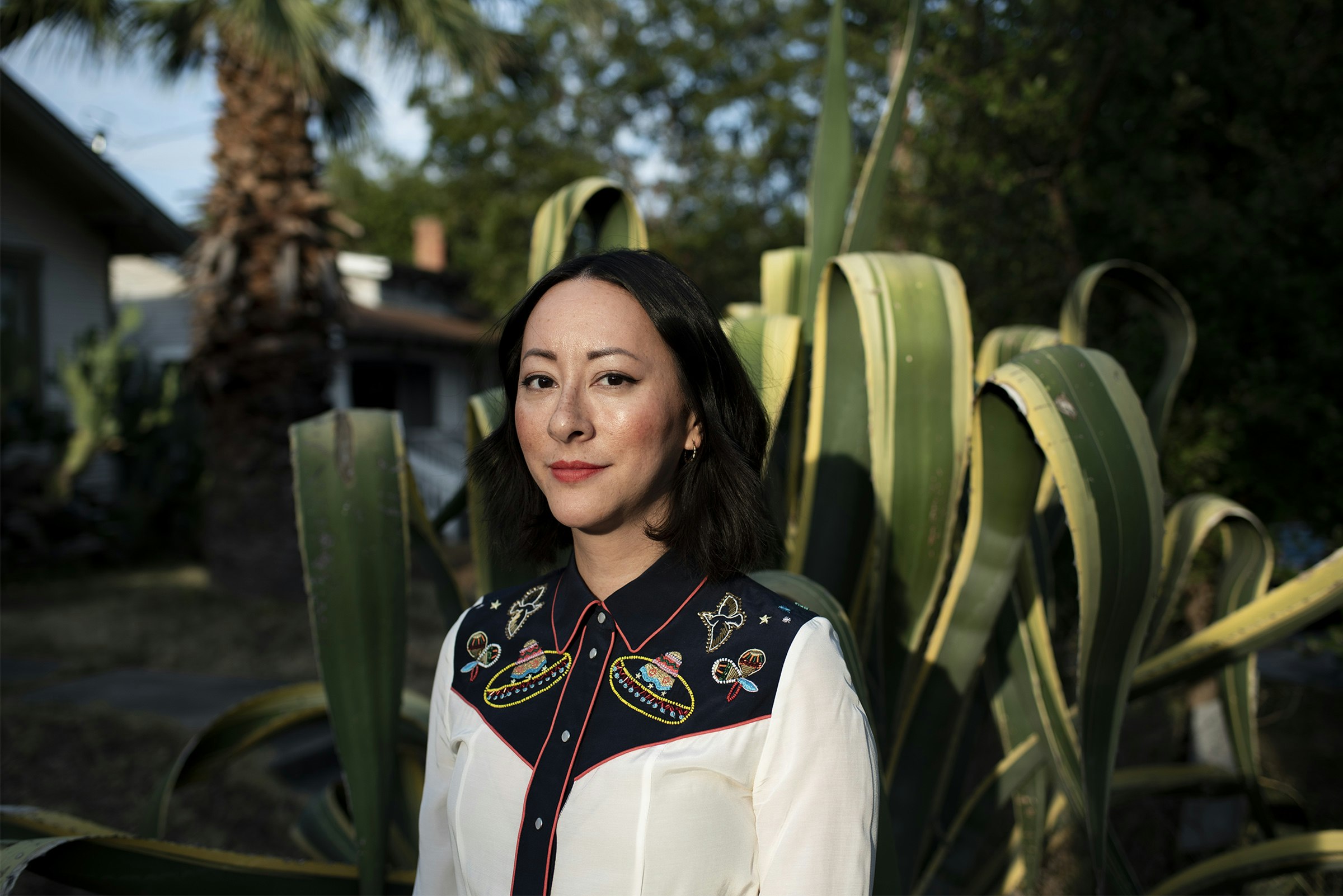 portrait of sun-illuminated person in a white and black embroidered western shirt posed infront of a large cactus in a yard with palm tree in background