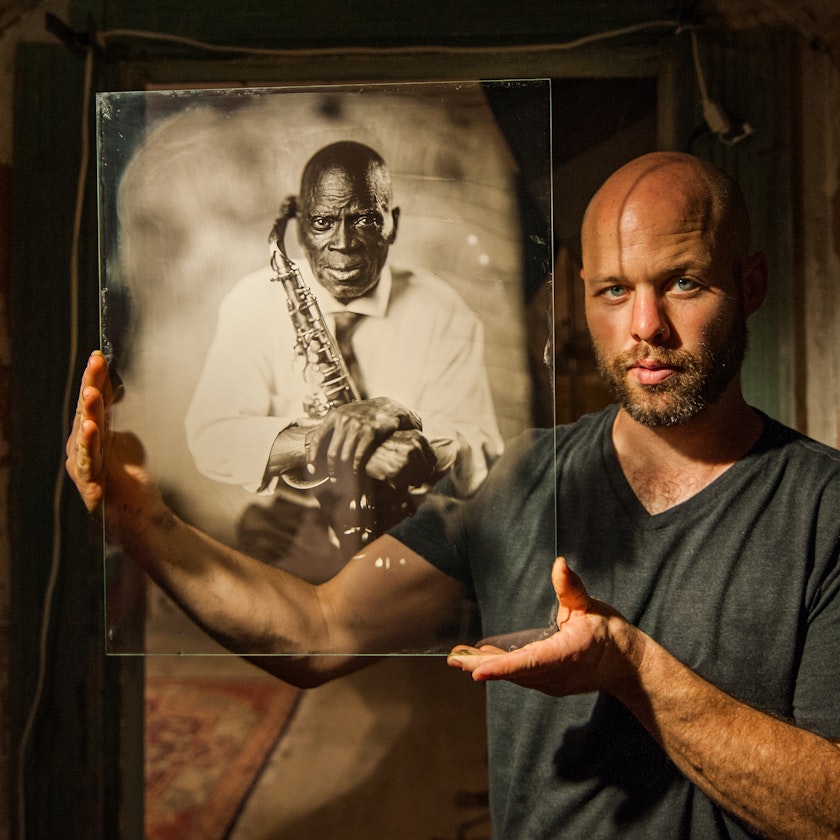 photographer with shaved head and beard holding up an ambrotype portrait of a person with a saxophone