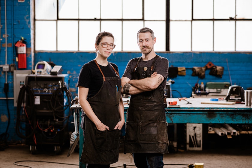 portrait of two blacksmiths wearing aprons posing in a shop with blue painted brick walls