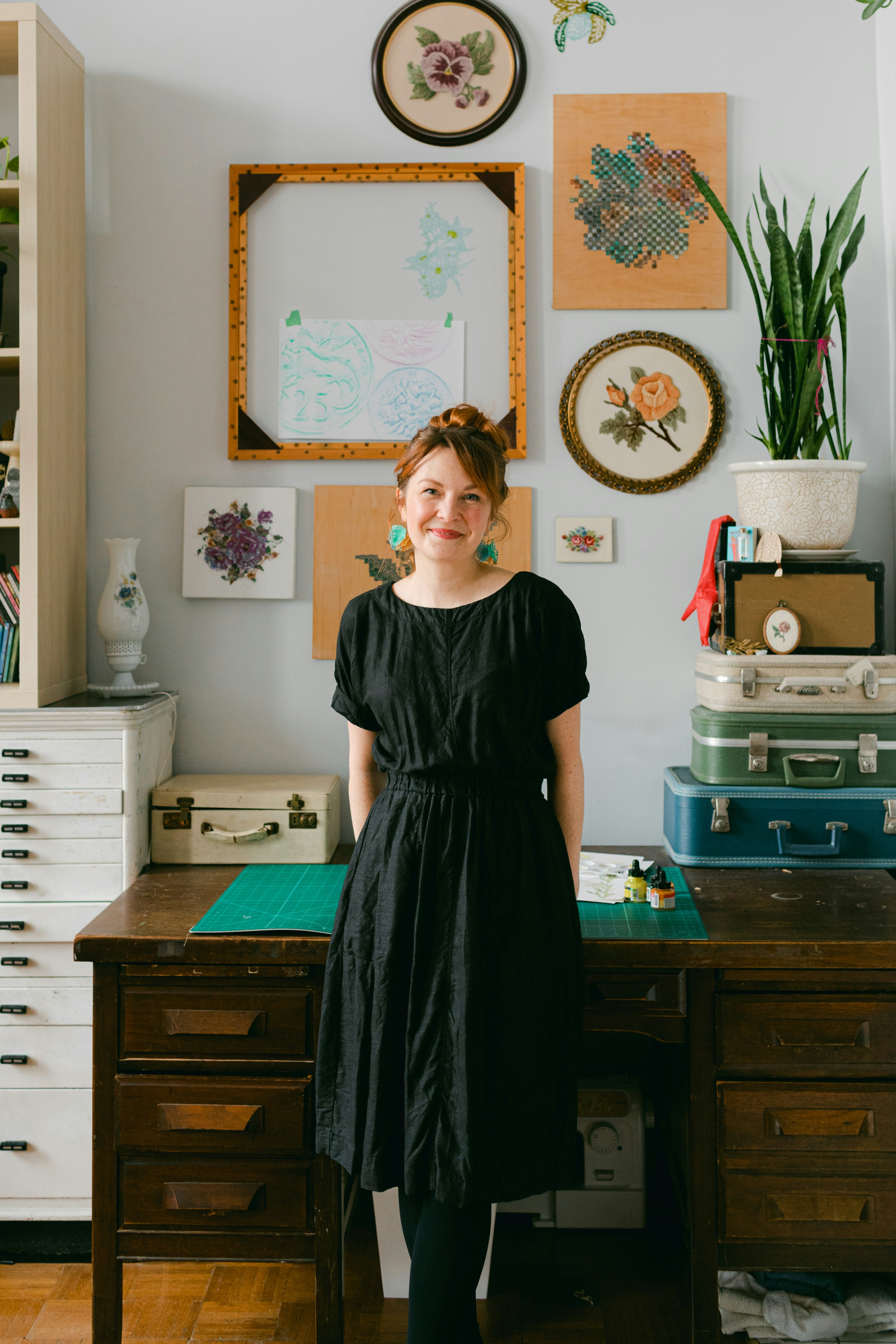 Portrait of Amanda McCavour posing against a desk in a studio with embroideries hanging on the walls