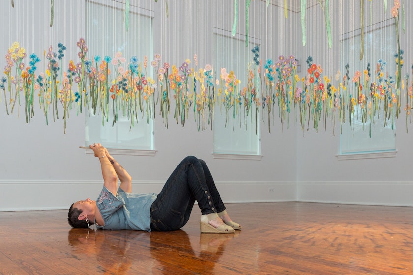 Person lying on a floor taking a photo of from beneath an installation of hanging embroidery ornaments