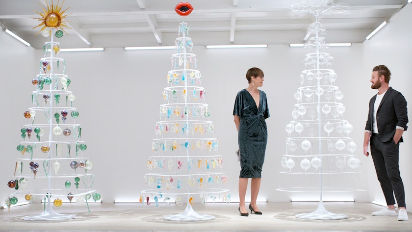 two glass critics reviewing hand-blown ornaments arranges on three christmas tree—like stands