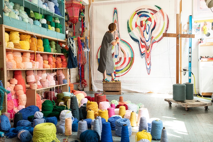 textile artist on a stool in a studio with shelves of yarn working on a colorful tufted wall hanging