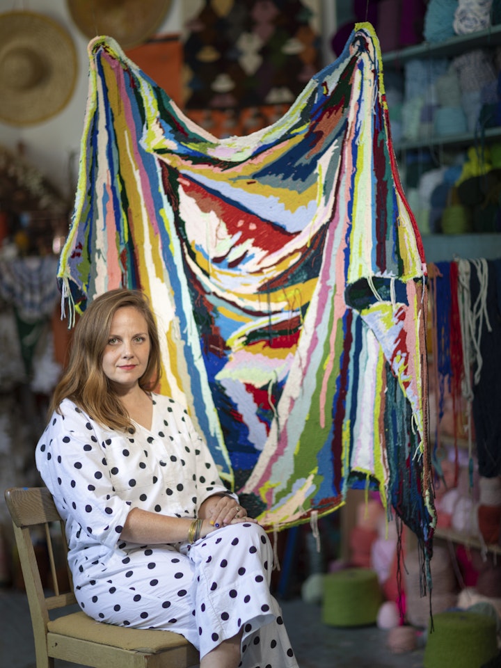 textile artist seated in studio beside colorful tufted wall hanging art