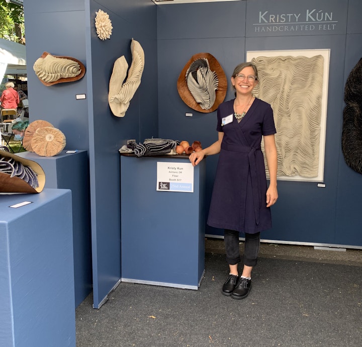 artist standing in exhibition booth with felt sculptures on display on blue walls