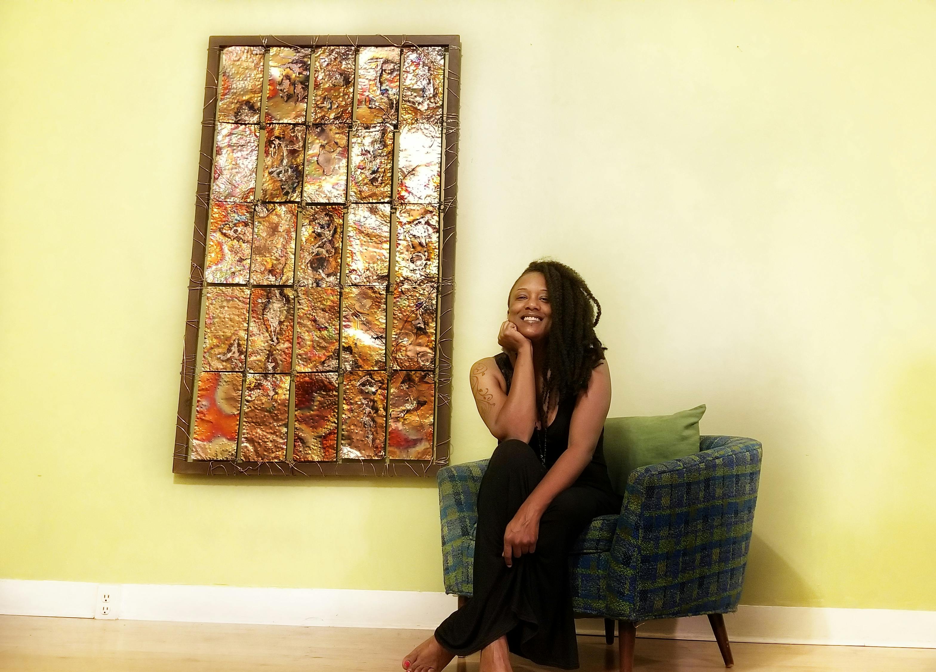 artist posing in green chair beside rectangular wallhanging featuring textured copper plates