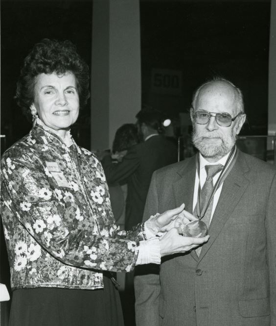 grayscale photo of a woman and man posingand smiling as the woman holds up a medal that the man is wearing around his neck