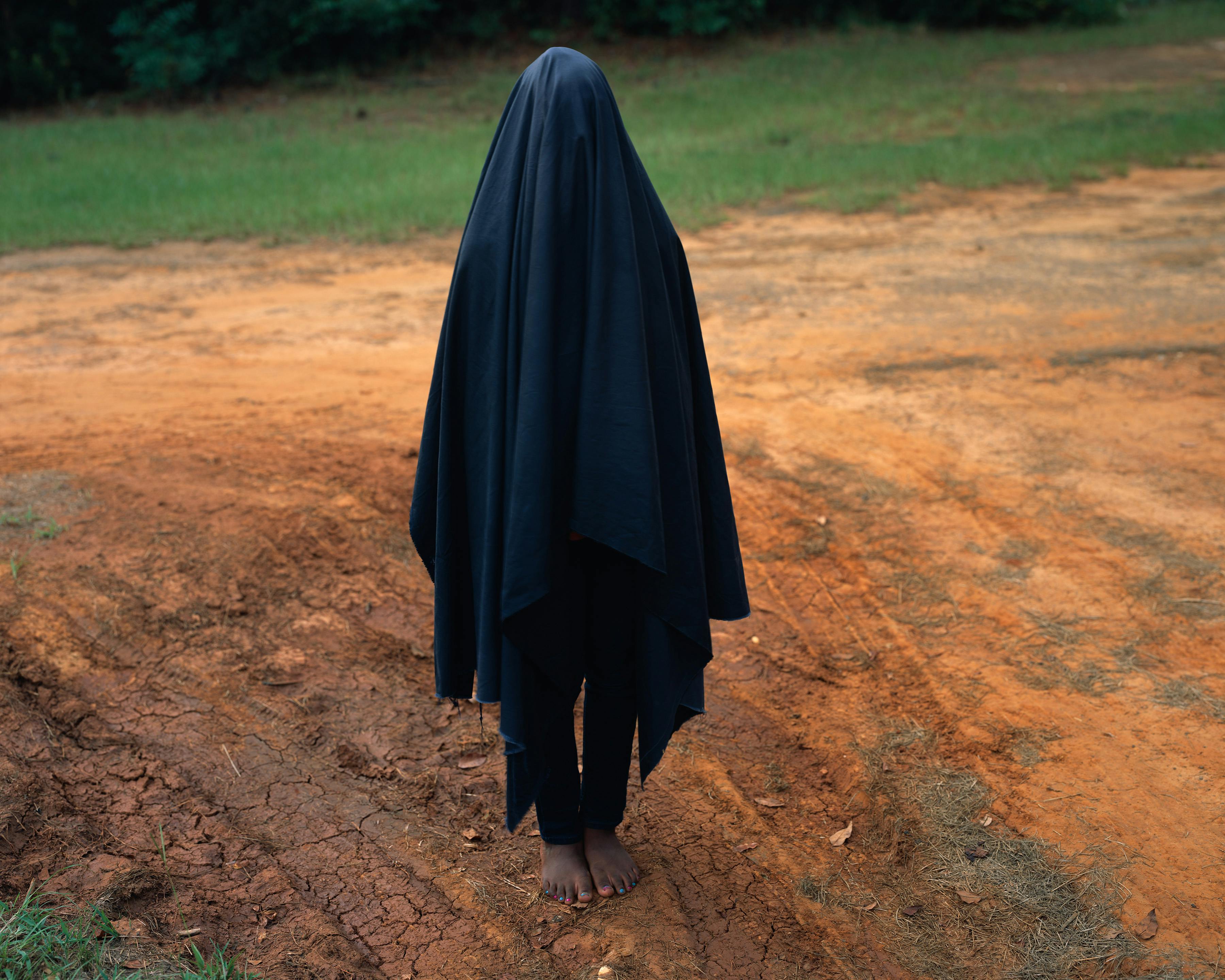 photograph of a person standing in a dirt road barefoot with a black veil covering their entire body