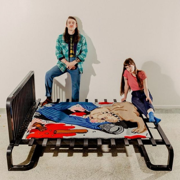 screenshot of an instagram post showing a two people posing with a black wooden bedframe that has a rug draped over it showing a bulldog chewing on a persons leg