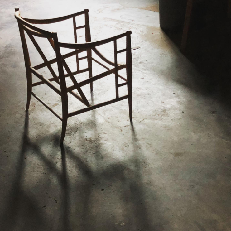 screenshot of an instagram post showing a silhouette of frame of a modern designed wooden chair on a concrete floor