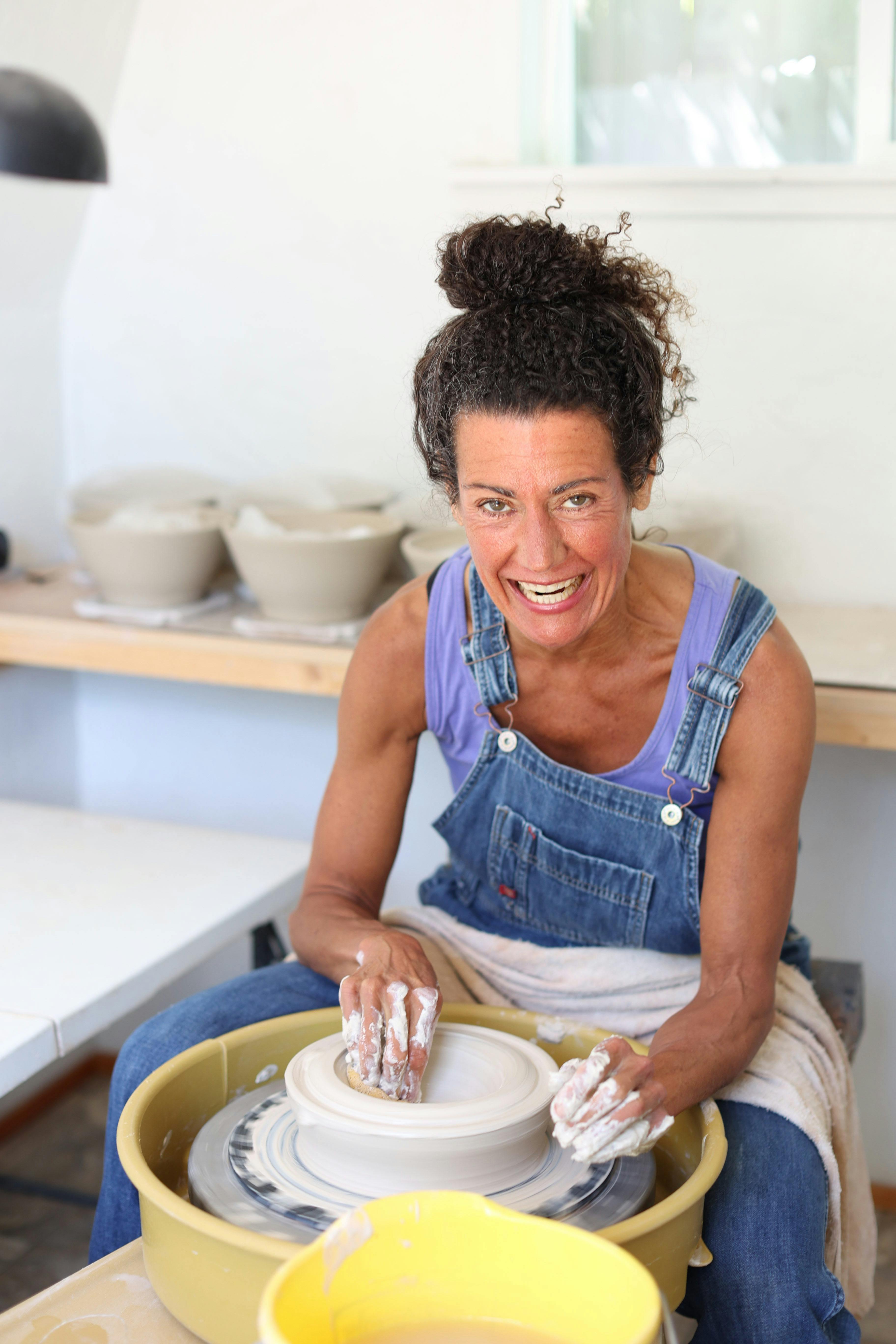 ceramic artist in purple tank top and overalls working on a pottery wheel and smiling