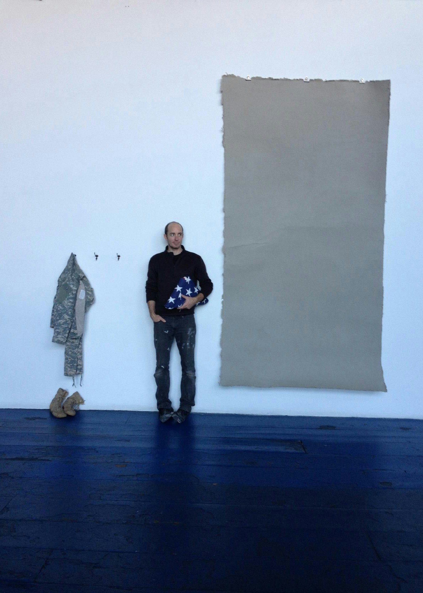 photo artist holding a folded american flag standing against a well in an exhibition space with a military uniform displayed on the left and a large sheet of tan olive paper displayed on the right