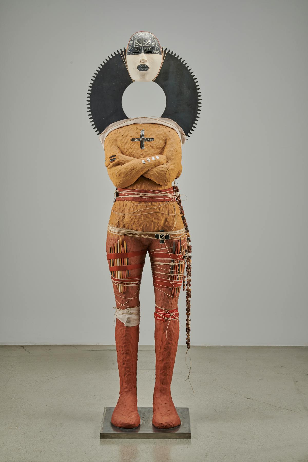 earthtone orange and red humanoid sculpture with arms folded and various accoutrements on legs and head suspended from body via a gray ring
