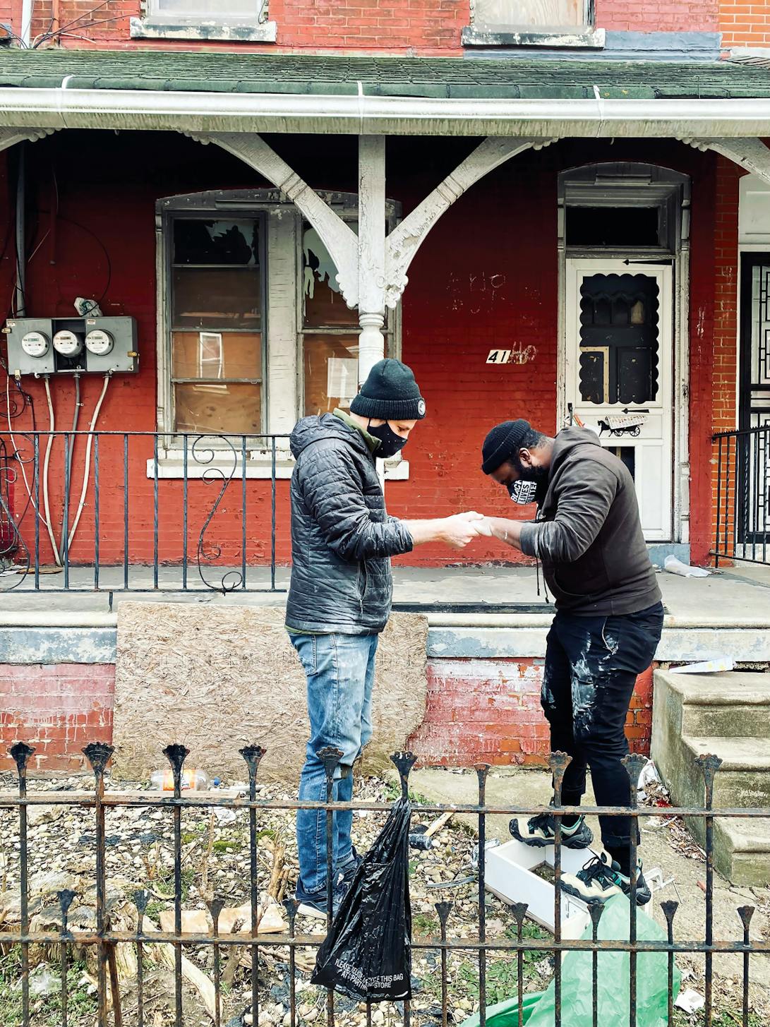 two people in face masks and jackets working together on a project in a yard outside a brick building