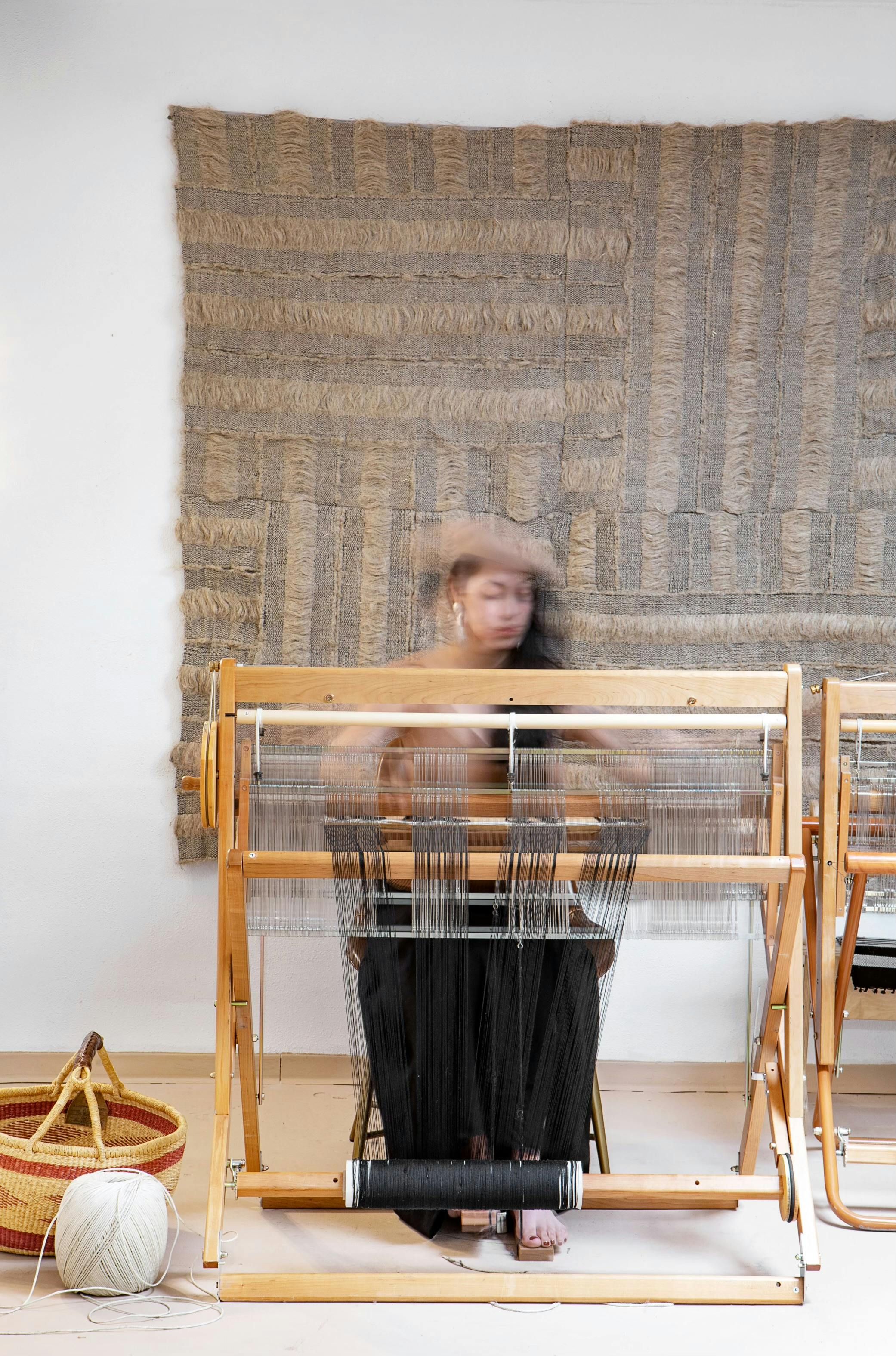 textile artist blurred in motion as they work at a loom. A finished beige piece hangs on the wall behind them